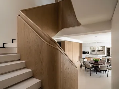 Modern interior view of a curved oak staircase with white risers overlooking a dining area featuring a round marble table and pendant lights.