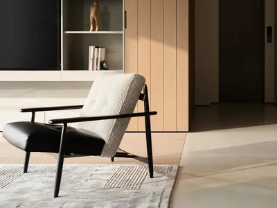 Modern living room featuring a black and white boucle armchair with black metal frame on a textured rug, positioned in front of built-in light wood and off-white cabinetry with open shelving and a large television.
