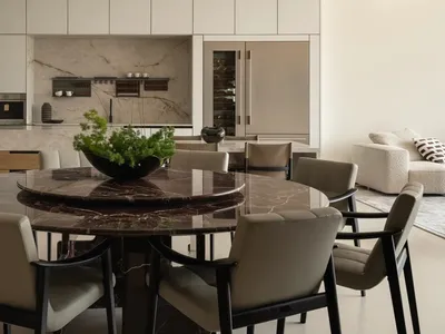 Modern minimalist dining area featuring a large round dark marble table, six beige upholstered chairs with black frames, and a striking white sculptural pendant lamp, with an open kitchen and living room in the background.