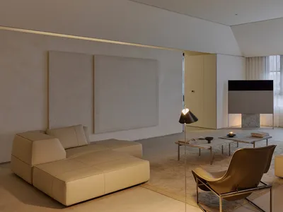Minimalist living room featuring a large beige modular sofa, brown leather lounge chair, floor lamp, and expansive micro-cement walls with warm ambient lighting.