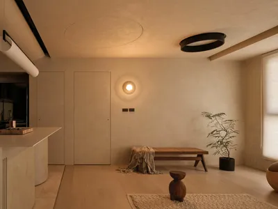 Modern minimalist open-plan kitchen and living room with warm beige microcement walls, light wood flooring, a central kitchen island with fluted detailing, wooden bench, potted tree, and circular ceiling lights.