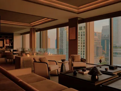 Modern luxury living room featuring a linear travertine fireplace, beige upholstered sofas, dark wood coffee tables, and floor-to-ceiling windows showcasing a dense city skyline at dusk.