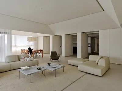 Spacious modern living room with beige leather sofas, micro-cement walls, concrete coffee tables, and a woman reading on the sofa in natural daylight.
