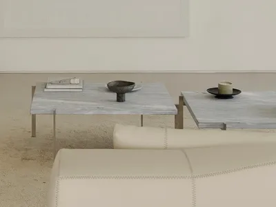 View of a minimalist living room featuring two square travertine coffee tables with thin metal legs, decorated with a dark stone bowl, stacked books, and a candle holder, set against a plain off-white wall and a cream-colored sofa in the foreground.