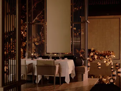 Elegant fine-dining restaurant interior featuring dark wooden lattice room dividers, white tablecloth dining tables, and golden leaf wall art under warm lighting.