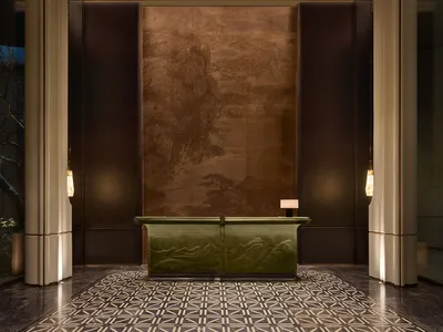 Luxurious hotel reception desk featuring a carved green marble console table against a large bronze panel with traditional Chinese landscape artwork, flanked by tall beige columns and geometric patterned marble floor.