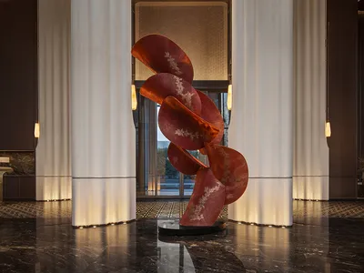 Luxurious hotel lobby featuring a large abstract red sculpture with gold detailing, flanked by tall white fluted marble columns on a polished dark marble floor.
