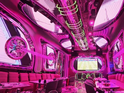 Futuristic nightclub lounge interior with pink and magenta neon lighting, circular acrylic mirrors, glowing green LED rings on the ceiling, and plush pink velvet seating.