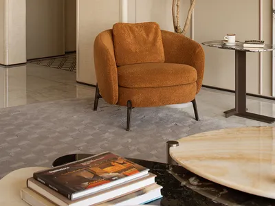 Modern living room corner featuring a burnt orange boucle armchair, tall potted tree, marble side table, and textured beige walls with wall sconce.