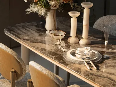 Elegant dining table with brown-veined Calacatta marble top, bouclé upholstered dining chairs, alabaster candle holders, and a large dried floral arrangement in a marble vase.