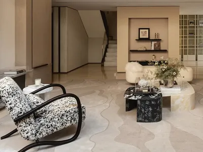 Modern double-height living room with a black and white patterned armchair, marble and terrazzo coffee tables, cream curved sofa, and a second-floor mezzanine with glass railing.