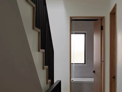 Modern minimalist hallway with black wooden staircase on the left and an open doorway leading to a bathroom with natural light at the end.