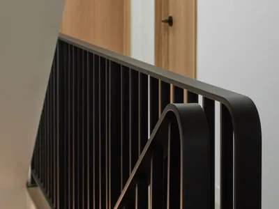 Close-up view of a modern black metal staircase railing with vertical bars and a curved handrail, paired with light wood treads and matching wood wall paneling.