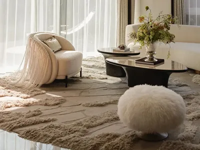 Bright modern luxury living room featuring a cream boucle fringed armchair, white fluffy ottoman, dark marble coffee table, and layered neutral rugs with large windows letting in natural light.