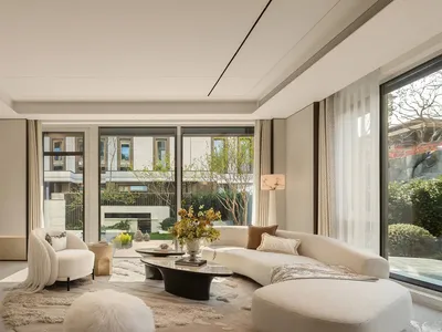 Modern minimalist living room featuring a large curved white boucle sofa, matching ottoman, white fur pouf, black coffee table, and floor-to-ceiling windows overlooking a lush garden.