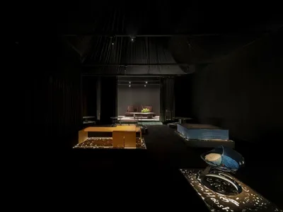 Dark contemporary lounge interior with black velvet curtains, low wooden seating, blue sculptural chair, and dramatic spotlighting on a raised platform.