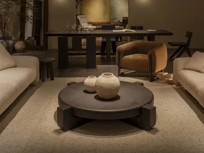 Modern minimalist living and dining area featuring a large round dark coffee table with textured ceramic vases, a mustard bouclé armchair, cream bouclé sofas, warm wall sconces, and abstract art in the background.
