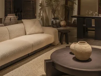 Modern organic living room featuring a cream boucle sofa, dark wood shutters, multiple ceramic vases, a round coffee table, and warm wall lighting.
