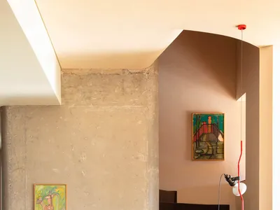 Modern brutalist interior featuring a dark concrete floating staircase, raw concrete pillar, warm wooden floor, and two colorful abstract paintings with a red pendant lamp.