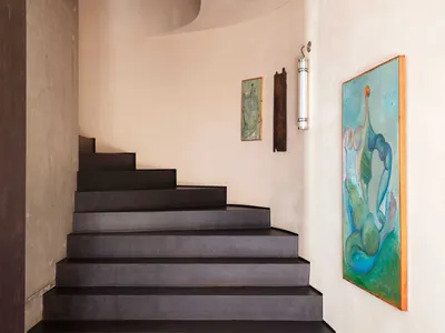 Modern interior view of a dark floating staircase with curved micro-cement walls, dark floor, and colorful abstract paintings on a light beige wall.