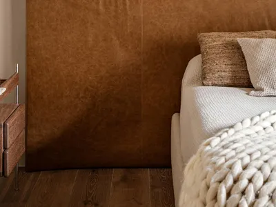 Close-up of a large brown leather upholstered headboard with visible texture, paired with cream knit bedding, a chunky white knitted throw, and a wooden nightstand on a dark wood floor.