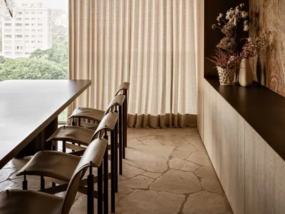 Modern minimalist dining area featuring a dark breakfast bar, four brown leather bar stools, irregular travertine stone floor, sheer beige curtains, wooden cabinetry, and a sculptural glass pendant light.