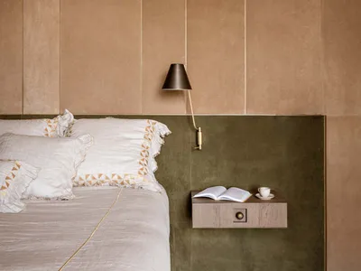 Modern minimalist bedroom featuring a warm beige upholstered wall, olive green velvet panel, floating wooden nightstand, brass wall lamp, and neutral linen bedding with geometric embroidery.
