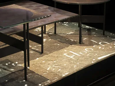 Two modern coffee tables, one rectangular with a glass top revealing a mosaic pattern and another round solid bronze top, placed on a thick textured rug with projected light patterns in a dark room.