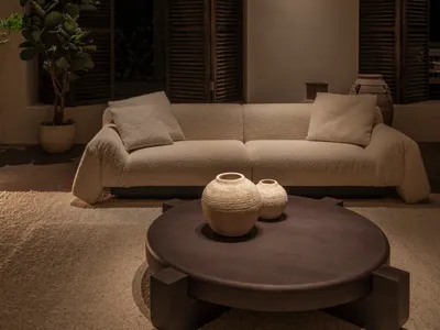Modern living room with a cream boucle sofa, round dark wood coffee table, textured ceramic vases, potted fiddle leaf fig, and dark wooden louvered shutters.