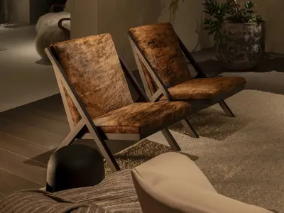 Two brown cowhide lounge chairs with dark wood frames placed on a textured rug in a warmly lit modern living room corner, accompanied by a large potted plant and wall sconce.