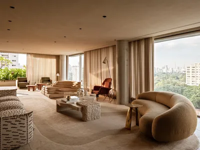Modern luxury living room featuring a large curved boucle sofa, travertine coffee tables, concrete structural pillar, and floor-to-ceiling windows overlooking a city skyline.