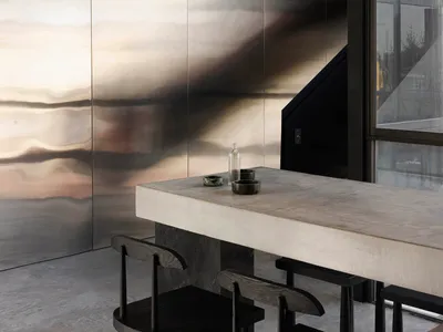 Minimalist dining area featuring a raw concrete island, dark wooden stools, and a large rippled stainless steel feature wall under a concrete ceiling.