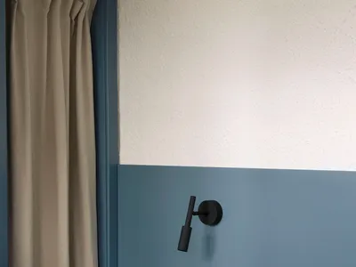 Modern bedroom corner featuring a two-tone blue wall, black wall sconce, round purple marble side table with books and a lamp, and a bed with neutral linen bedding.