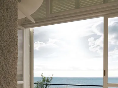 Modern coastal living space with blue and white patterned terrazzo floor tiles, white marble coffee table, wooden zigzag chair, and large open doors revealing a panoramic ocean view under a bright blue sky.