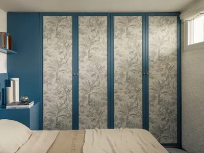 Modern bedroom with deep blue walls, a built-in wardrobe covered in grey tropical leaf wallpaper, a floating blue shelf with books, and a bed with neutral linen bedding.