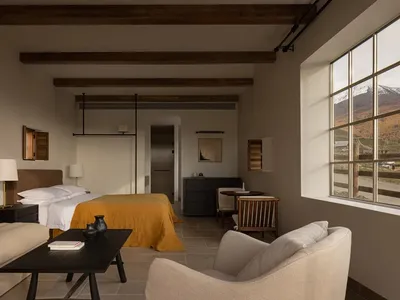 Modern mountain lodge bedroom featuring exposed dark wood ceiling beams, a bed with mustard yellow throw, beige sofa, dark coffee table, and large window overlooking snowy mountain landscape.