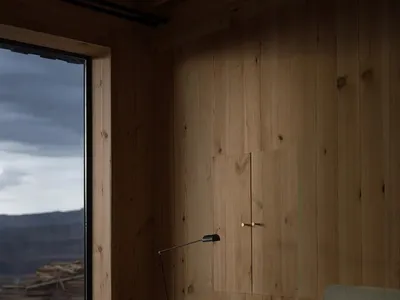 Cozy mountain cabin bedroom featuring light wood panel walls, a dark wood writing desk, woven rattan chair, and large window overlooking a dramatic landscape.