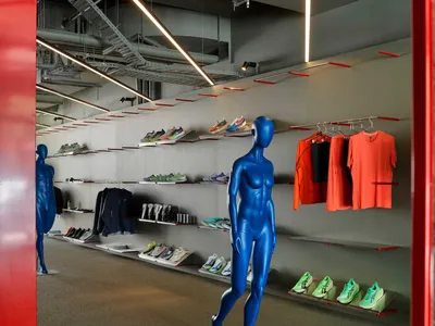 Interior view of a contemporary sportswear store featuring bright blue glossy mannequins, wall-mounted display shelves with colorful sneakers, hanging athletic apparel, and dramatic linear LED lighting.