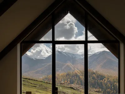 Interior view of a modern mountain lounge featuring a large black-framed gable window overlooking a vast mountain range with autumn foliage and snow-capped peaks, with minimalist wooden furniture in the foreground.