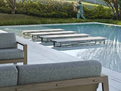 Modern infinity pool with teak and fabric lounge chairs on a stone deck, a person tending to plants in the background with forested hills beyond.