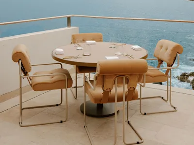 Round outdoor dining table with four mustard-ochre upholstered chairs on a stone balcony overlooking the Adriatic Sea and the old town of Dubrovnik, Croatia.