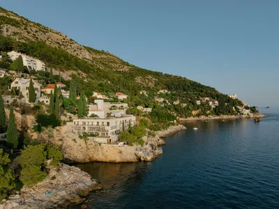 A modern white cliffside hotel built into rocky terraces overlooking the deep blue Adriatic Sea, with people sunbathing on the rocks below and green hills rising behind.