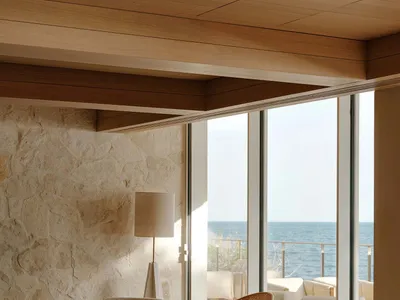 Modern coastal living room featuring a large travertine stone wall, two burnt orange lounge chairs, neutral sofa, glass coffee table, and panoramic ocean view through floor-to-ceiling windows.