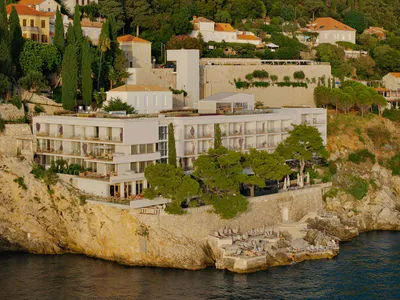Modern white luxury hotel built into a rocky cliffside with multiple terraces, stone retaining walls, and direct access to the sea below.