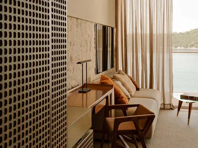 Modern coastal living room featuring a walnut console, wooden armchair, beige sofa with orange cushions, perforated wooden screen, and large window overlooking the sea.