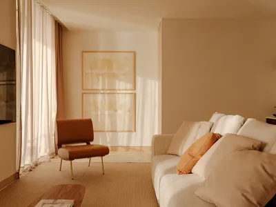 Minimalist living room featuring a cream boucle sofa with terracotta and beige cushions, a burnt orange leather accent chair with brass legs, two abstract watercolor artworks in wooden frames, sheer linen curtains, and warm natural sunlight creating soft shadows on beige walls.