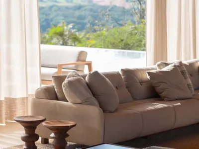 Modern neutral living room featuring a beige modular sofa, wooden accent tables, textured jute rug, and large windows overlooking green hills.