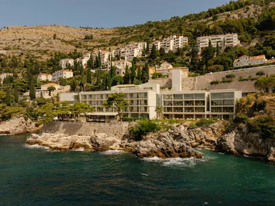 A contemporary white hotel with extensive glass windows built directly on rocky cliffs above the deep blue sea, with a hillside town in the background under a clear sky.