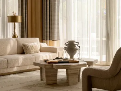Bright minimalist living room featuring a cream boucle sofa, travertine coffee table, sculptural white vase, and sheer curtains with warm neutral tones.