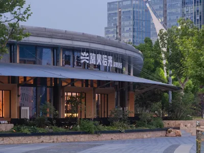 現代弧形餐廳外觀，石材基座、大面積玻璃窗與發光招牌，在黃昏時分，周圍環繞樹木與城市建築。